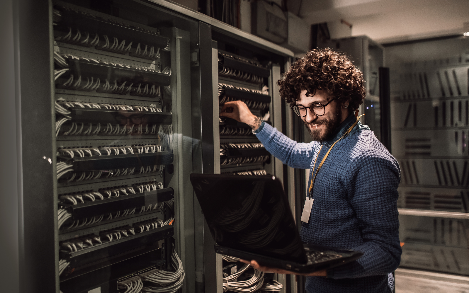 A technician with a laptop works on a server rack with numerous cables in a data center. This highlights Hybrid Cloud Computing for enterprise, enabling efficient management and scalability for business needs in a modern IT environment.