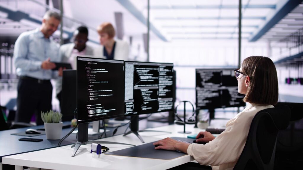 A person working at a desk with multiple monitors displaying code, in a modern office with colleagues collaborating in the background.