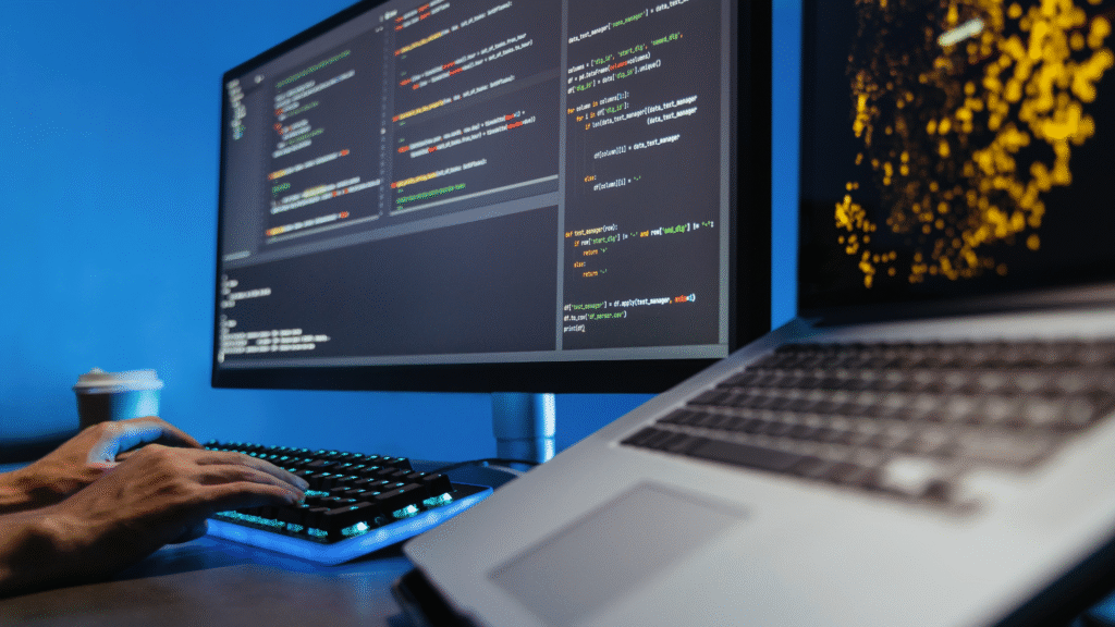 A person typing on a keyboard with a monitor displaying code and a laptop nearby, set against a blue background with a coffee cup, focusing on programming.