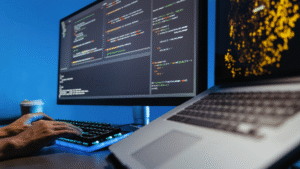 A person typing on a keyboard with a monitor displaying code and a laptop nearby, set against a blue background with a coffee cup, focusing on programming.