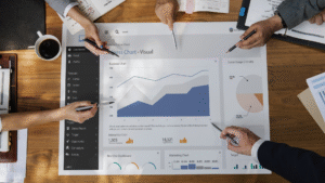 People analyzing a large business chart visual with graphs and data on a wooden table, surrounded by coffee cups, notebooks, and pens, focusing on business analytics.