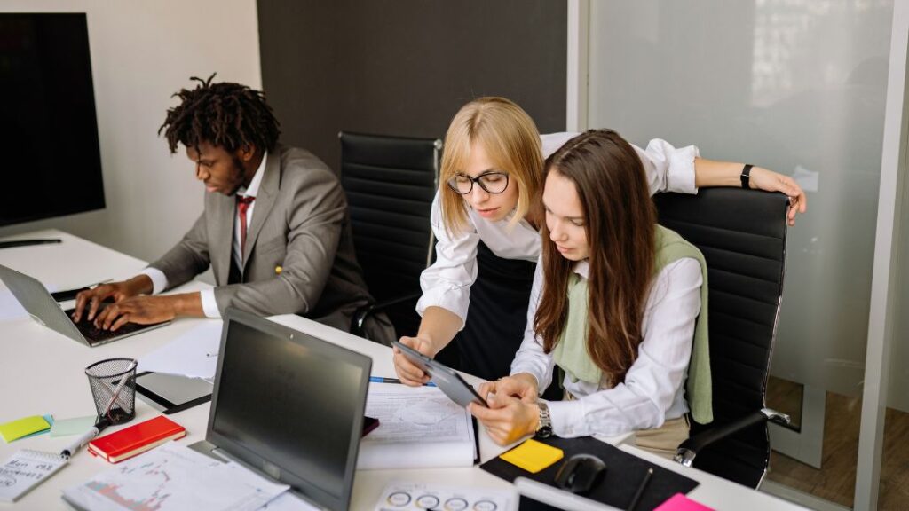 Data Cleansing for BI Enterprise Strategies, Tools & Best Practices. Blonde woman with glasses leaning over to show a tablet to a brunette colleague at a desk, while a man in a suit works on a laptop in the background.